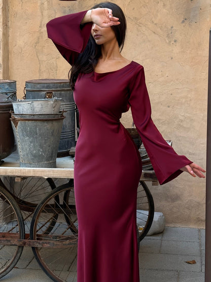 Donna in abito lungo bordeaux con maniche a campana, posa davanti a vecchi secchi di metallo e ruote di carro, sfondo rustico.