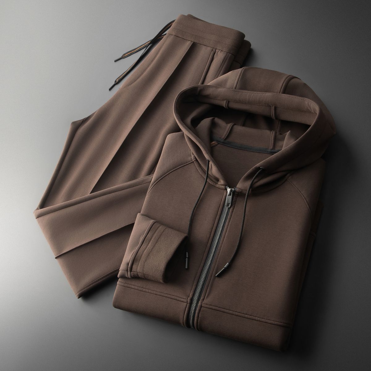 Tuta sportiva marrone in cotone, composta da felpa con cappuccio e pantaloni, su sfondo grigio. Abbigliamento casual, moda uomo, comfort.