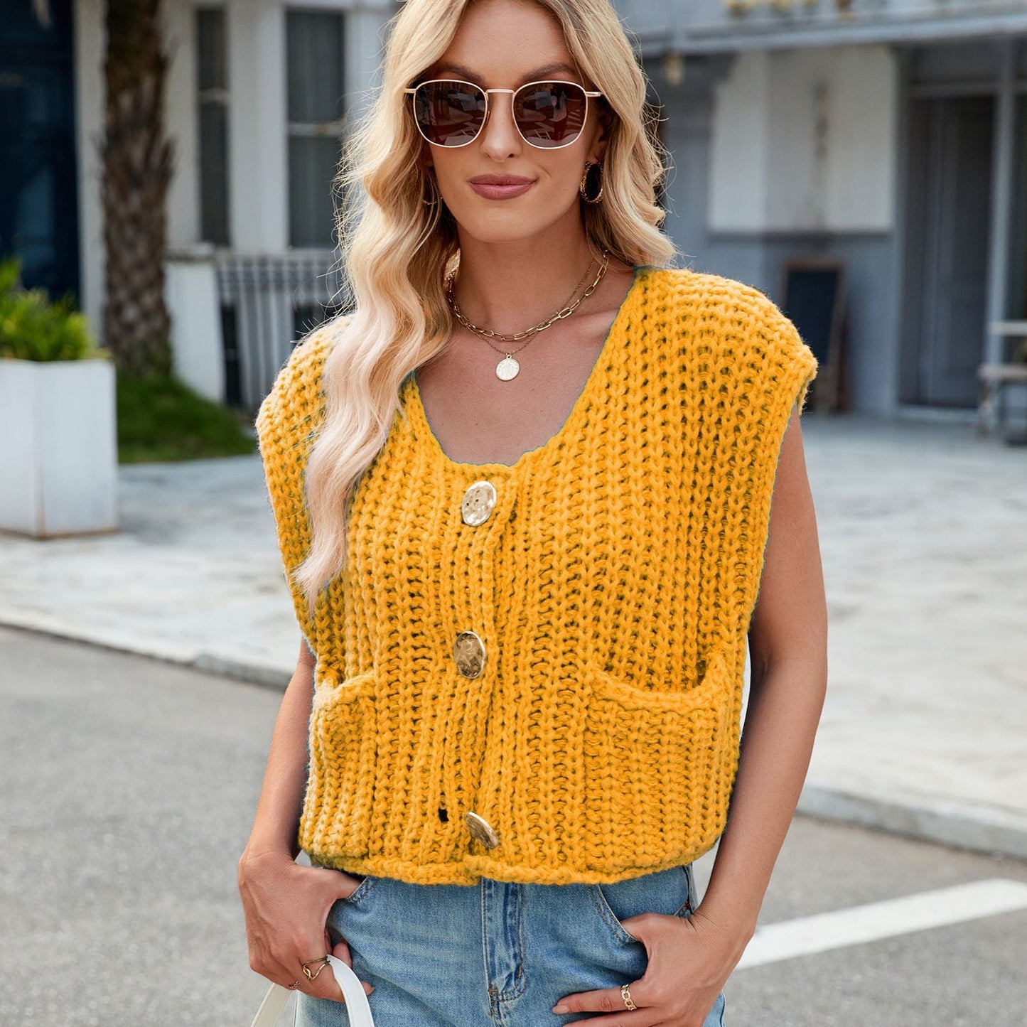 Donna con occhiali da sole indossa un gilet giallo lavorato a maglia con bottoni grandi e tasche, su strada urbana. Moda autunnale casual chic.