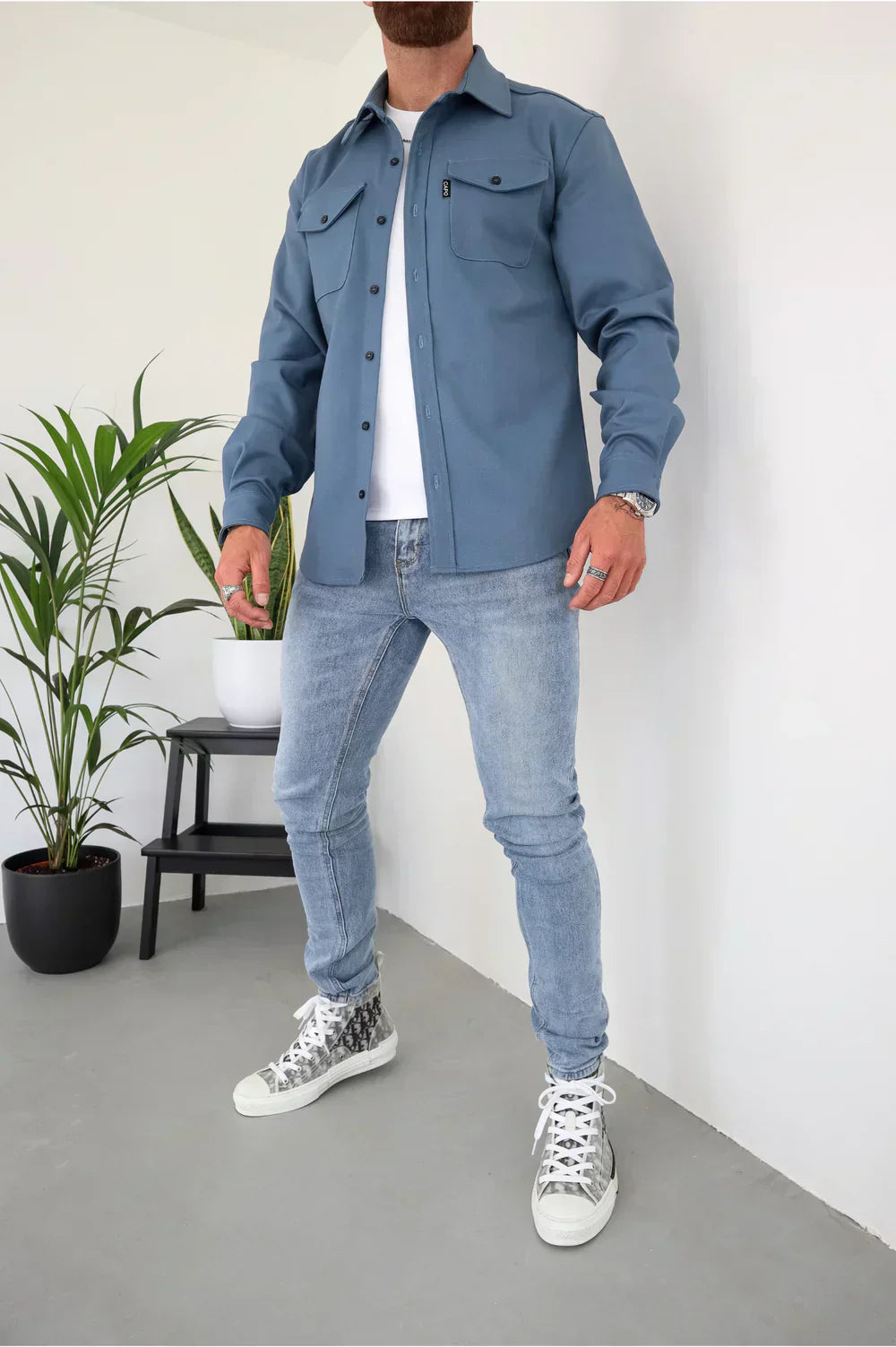 Uomo in camicia blu e jeans chiari, scarpe da ginnastica bianche, accanto a pianta in vaso e sedia nera, moda casual maschile moderna.