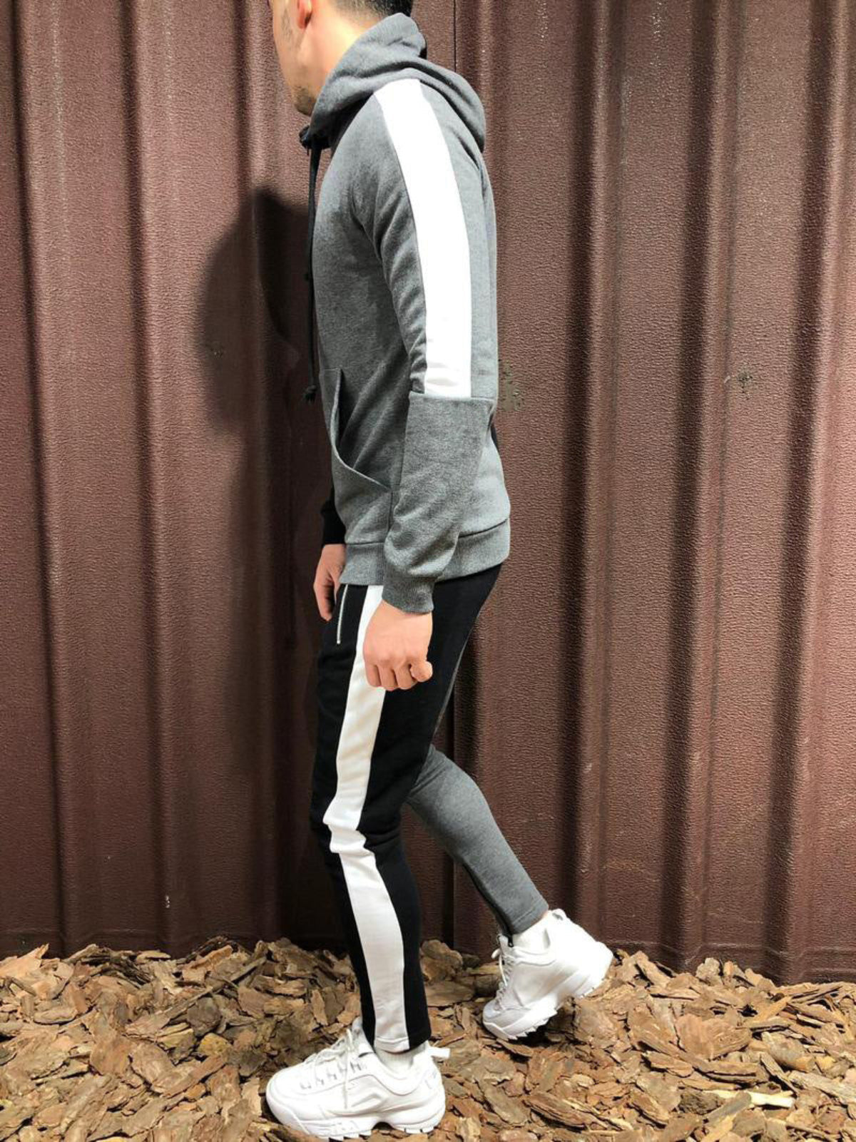 Uomo in tuta sportiva grigia con strisce bianche, scarpe da ginnastica bianche, su sfondo di lamiera marrone e foglie secche.