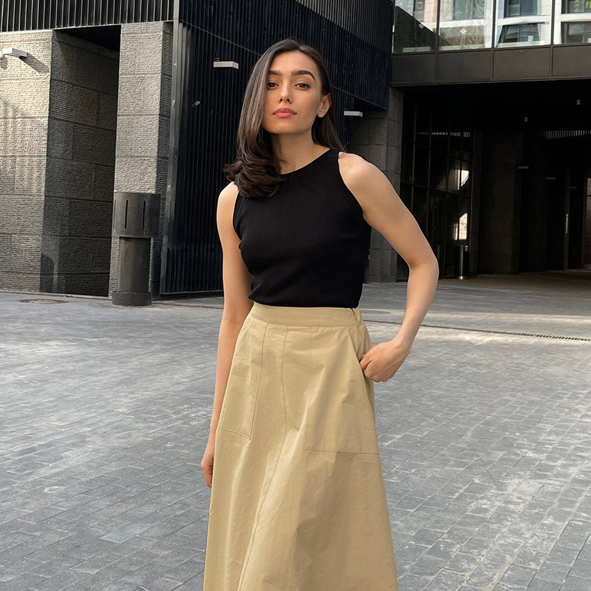 Donna con top nero e gonna beige in posa su strada urbana, architettura moderna sullo sfondo. Moda casual elegante, look estivo chic.