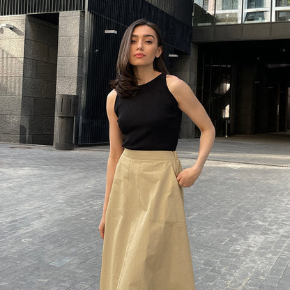 Donna con top nero e gonna beige in posa su strada urbana, architettura moderna sullo sfondo. Moda casual elegante, look estivo chic.