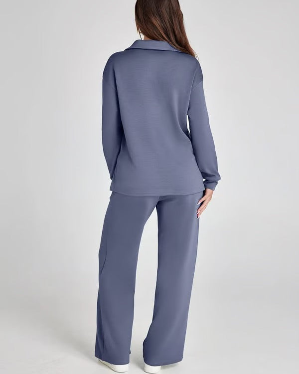 Donna di spalle in completo loungewear blu, maglia a maniche lunghe e pantaloni larghi, su sfondo grigio. Moda casual, abbigliamento comodo.