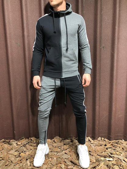 Tuta sportiva uomo bicolore nero e grigio, con cappuccio e pantaloni abbinati, indossata con scarpe bianche su sfondo marrone.
