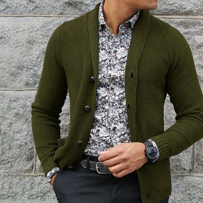 Uomo con cardigan verde oliva, camicia a fiori bianca e nera, orologio nero, pantaloni scuri. Moda maschile elegante e casual.
