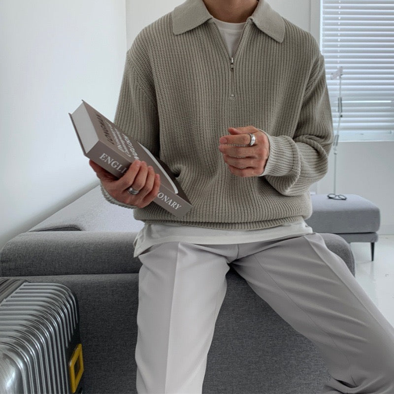 Uomo seduto su divano grigio con maglione beige, pantaloni chiari, tiene un libro "English Dictionary". Interno moderno, valigia metallica accanto.
