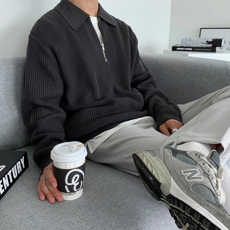 Uomo seduto su divano grigio indossa maglione scuro e pantaloni chiari, tiene caffè da asporto. Scarpe sportive grigie New Balance in primo piano.