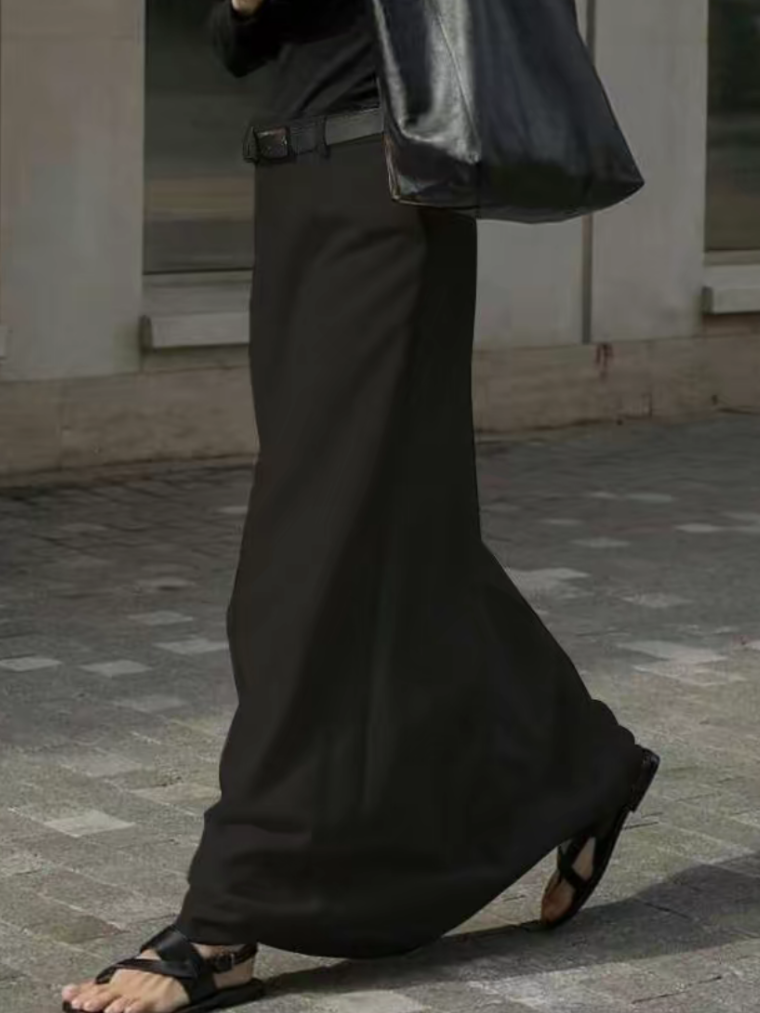 Uomo con gonna lunga nera, sandali e borsa in pelle nera, cammina su marciapiede. Moda maschile, abbigliamento unisex, stile urbano.