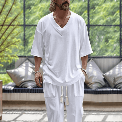 Uomo con abbigliamento casual bianco, maglietta e pantaloni larghi, in un ambiente moderno con divano e piante, moda estiva maschile.