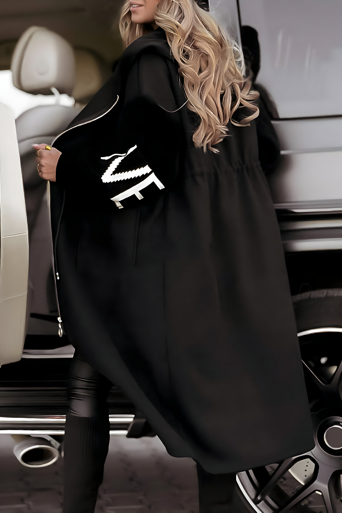 Donna con cappotto nero elegante, dettagli bianchi, capelli biondi ondulati, accanto a un'auto di lusso. Moda invernale, stile sofisticato.