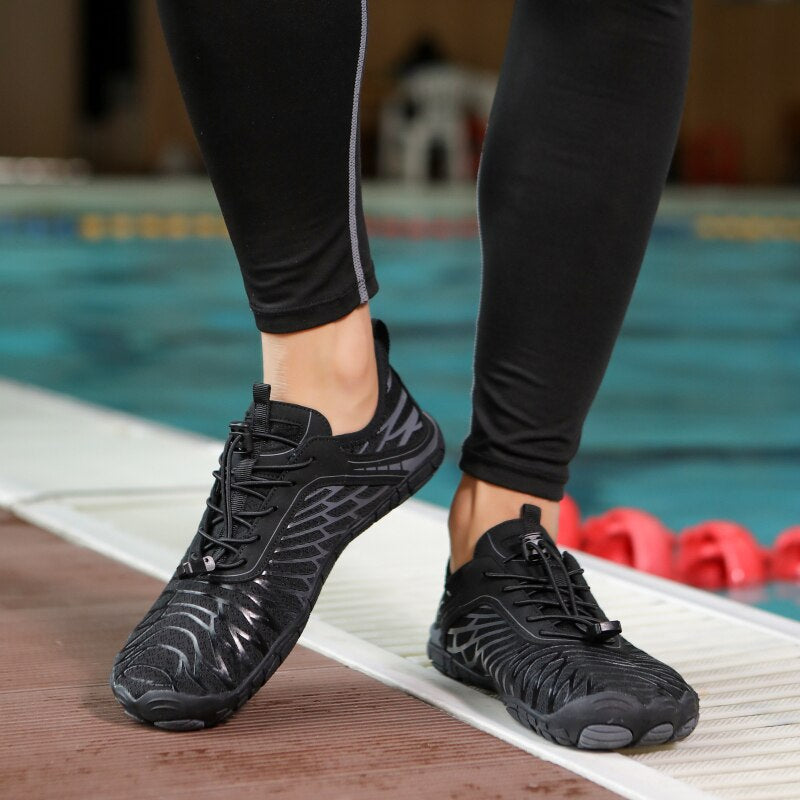 Scarpe da acqua nere unisex, design sportivo, suola antiscivolo, indossate vicino a una piscina. Ideali per nuoto e attività acquatiche.