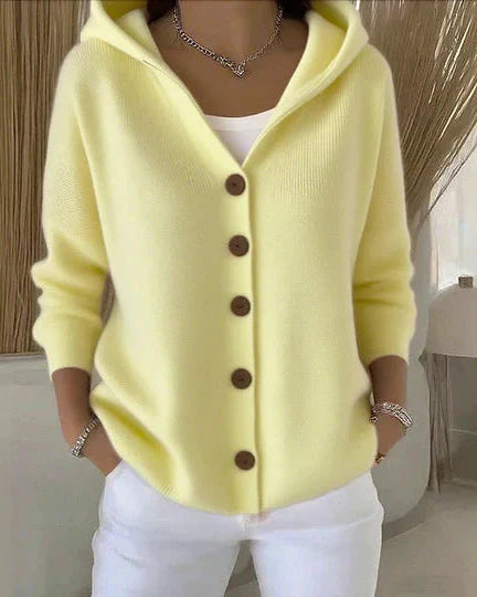 Donna con cardigan giallo pastello con cappuccio e bottoni marroni, indossa pantaloni bianchi. Stile casual elegante, moda autunnale femminile.