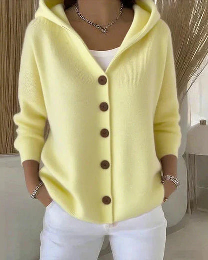 Donna con cardigan giallo pastello con cappuccio e bottoni marroni, indossa pantaloni bianchi. Stile casual elegante, moda autunnale femminile.