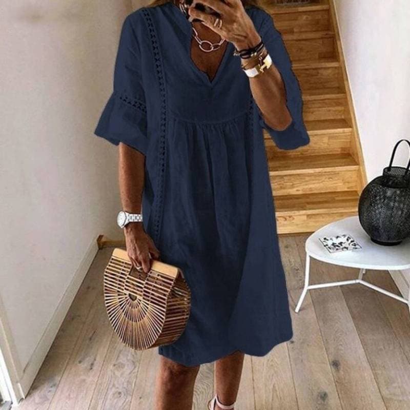 Donna con abito blu scuro estivo, borsa in legno a mezzaluna, orologio bianco, collana e bracciali, in piedi su pavimento in legno vicino a scale.