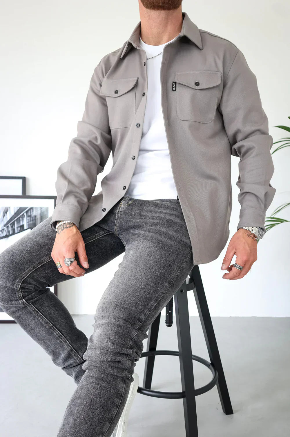 Uomo seduto su sgabello indossa camicia grigia con tasche, jeans grigi e maglietta bianca. Moda casual maschile, stile urbano elegante.