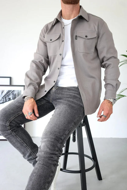 Uomo seduto su sgabello indossa camicia grigia con tasche, jeans grigi e maglietta bianca. Moda casual maschile, stile urbano elegante.