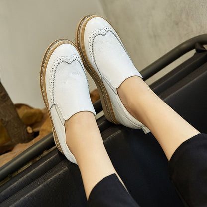 Scarpe slip-on bianche da donna in pelle, con dettagli traforati e suola in gomma marrone, indossate su un divano nero. Moda calzature eleganti.