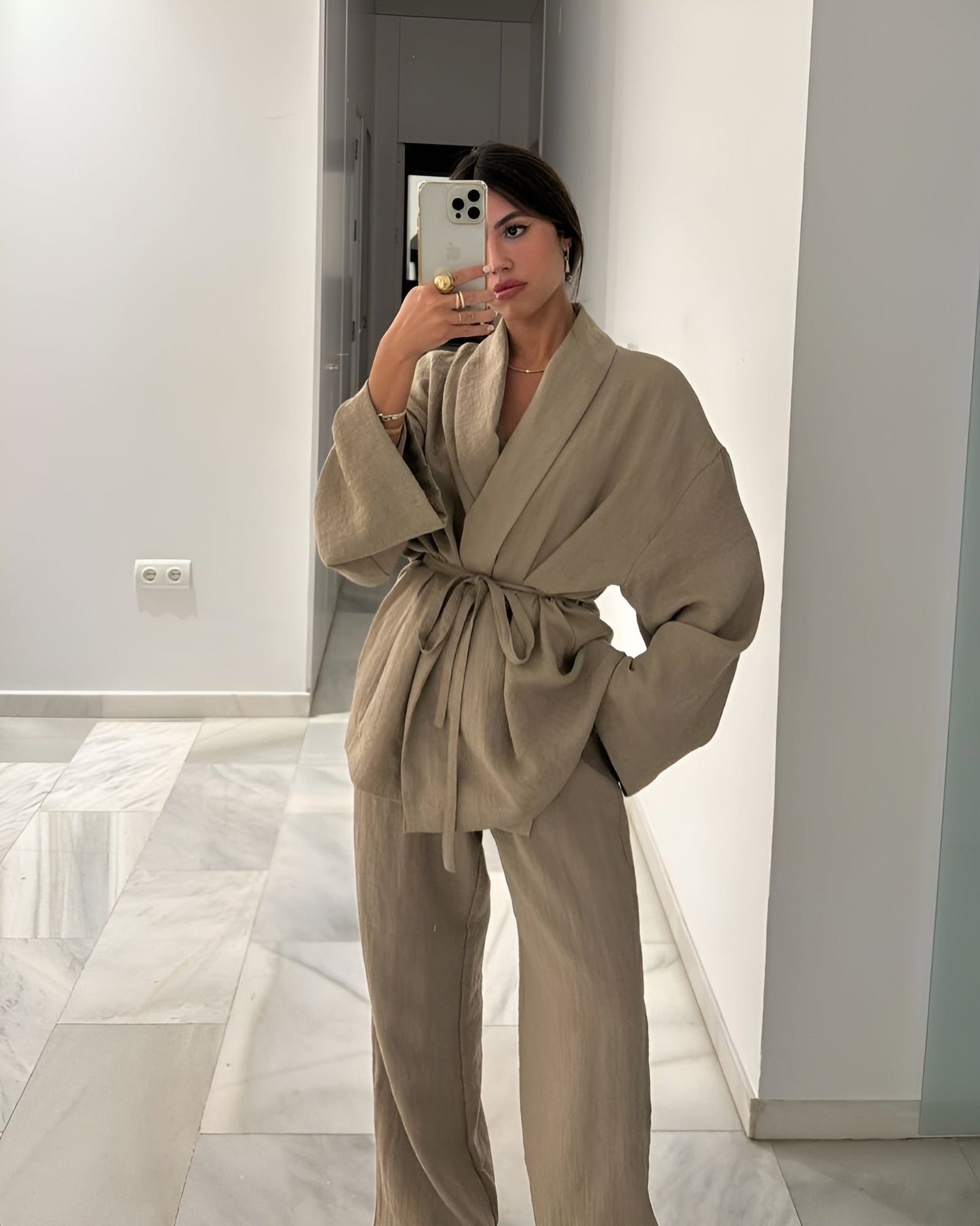 Donna in abito kimono beige scatta selfie con iPhone in corridoio moderno. Moda minimalista, outfit elegante, tendenze abbigliamento 2023.