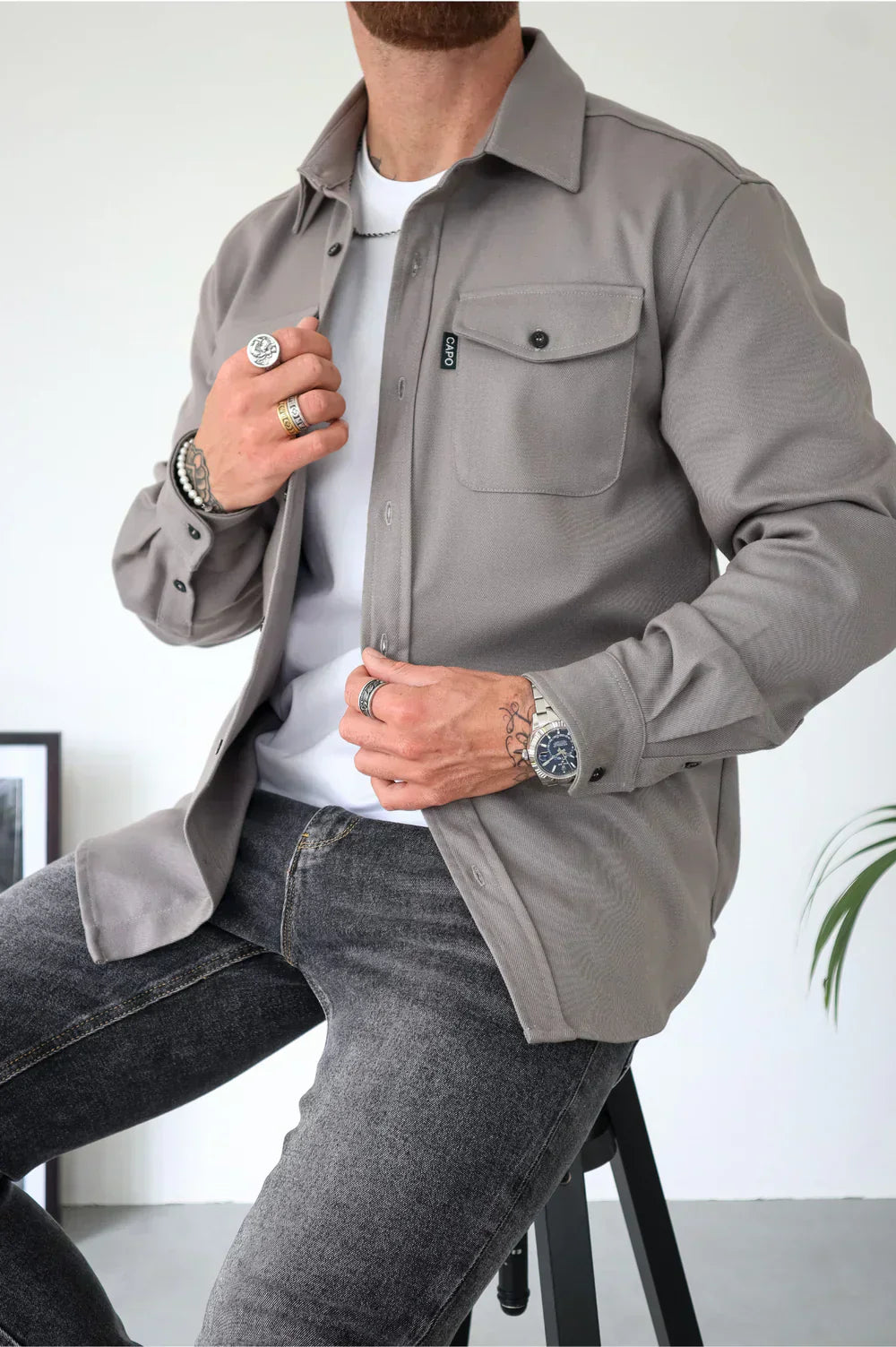 Uomo con camicia grigia casual, seduto su sgabello, indossa jeans scuri, orologio e anelli. Stile moderno e minimalista, moda maschile.