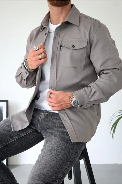 Uomo con camicia grigia casual, seduto su sgabello, indossa jeans scuri, orologio e anelli. Stile moderno e minimalista, moda maschile.