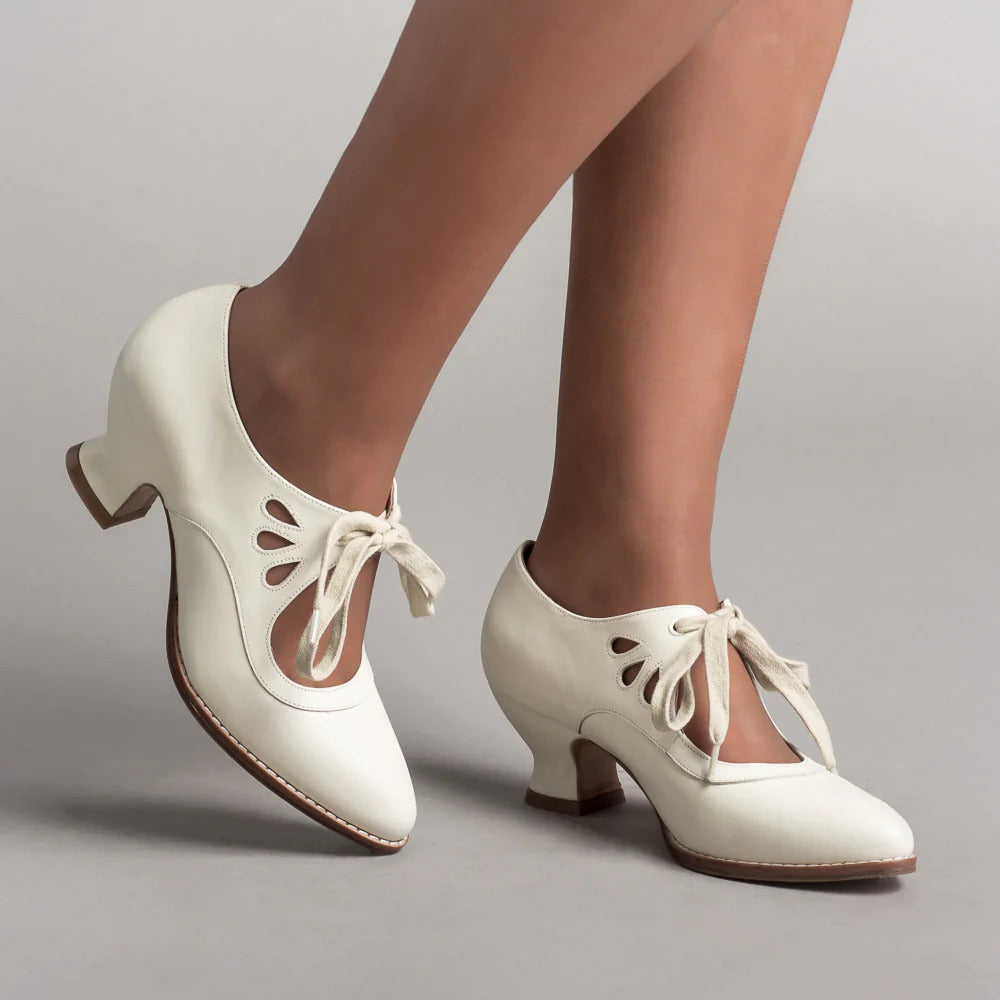 Scarpe da ballo vintage bianche con tacco medio, lacci decorativi e dettagli traforati, indossate su sfondo grigio. Eleganza retrò per danza.