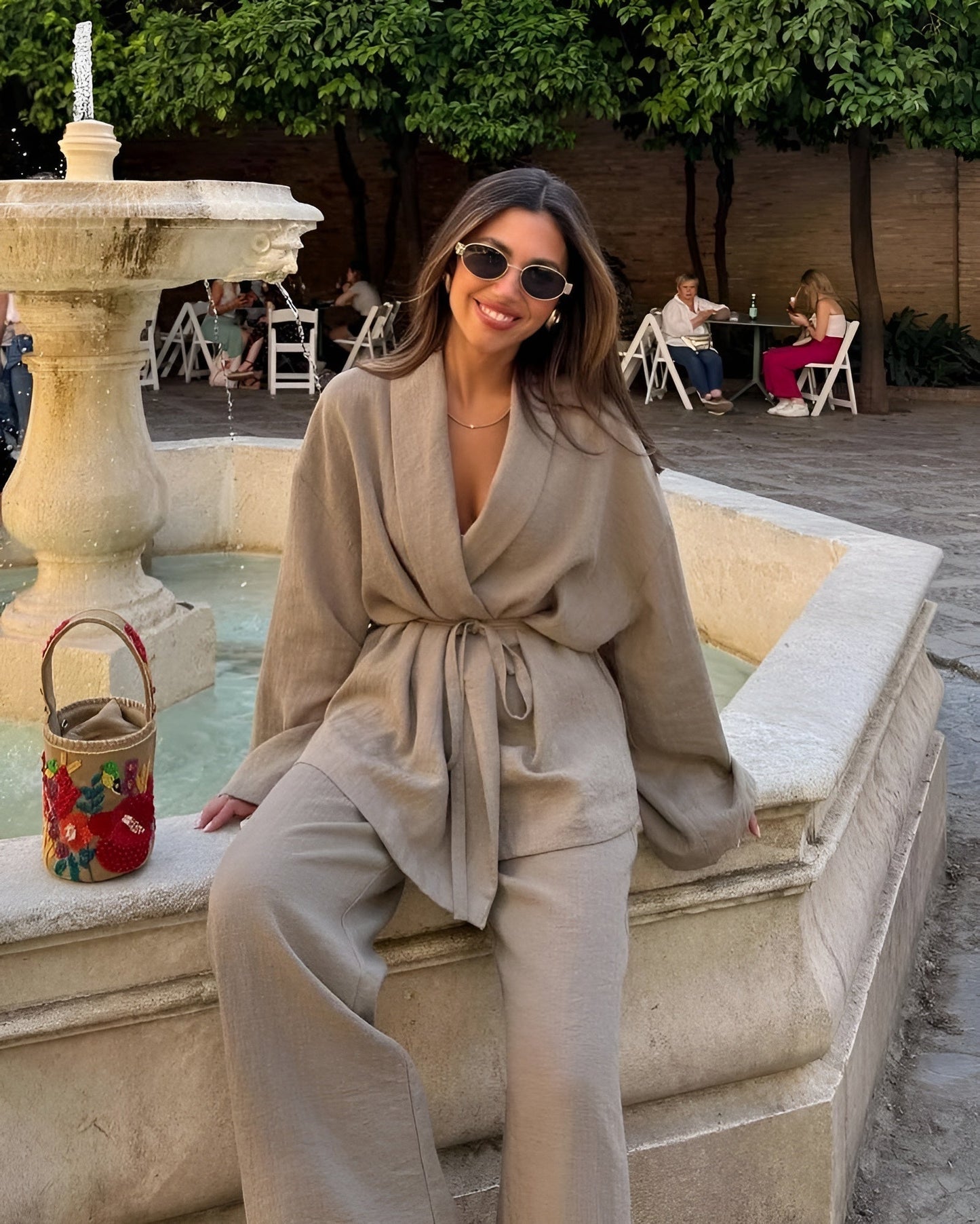Donna sorridente con occhiali da sole, indossa abito beige elegante, seduta su una fontana in pietra. Borsa colorata accanto, ambiente esterno rilassante.