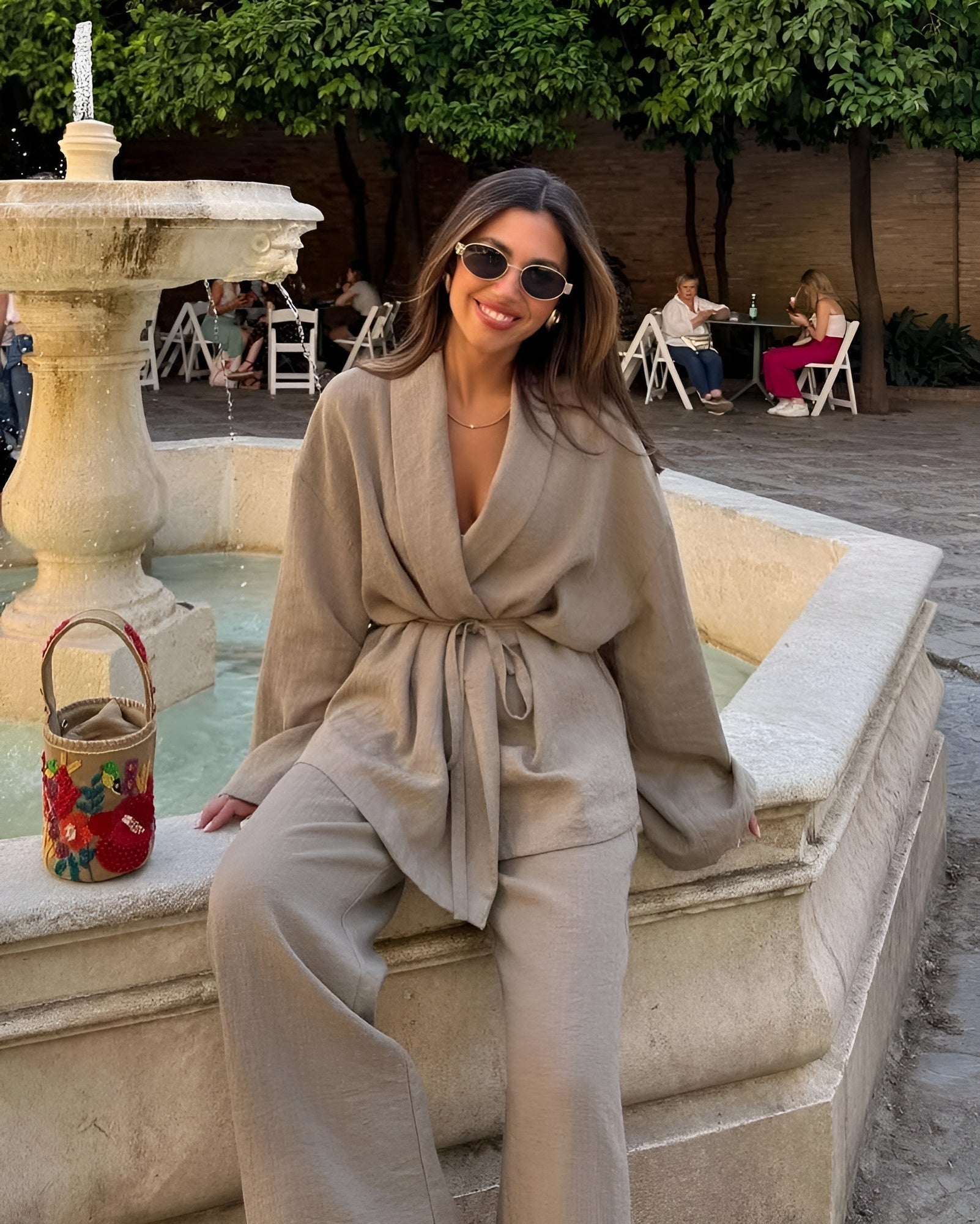Donna sorridente con occhiali da sole, indossa abito beige elegante, seduta su una fontana in pietra. Borsa colorata accanto, ambiente esterno rilassante.