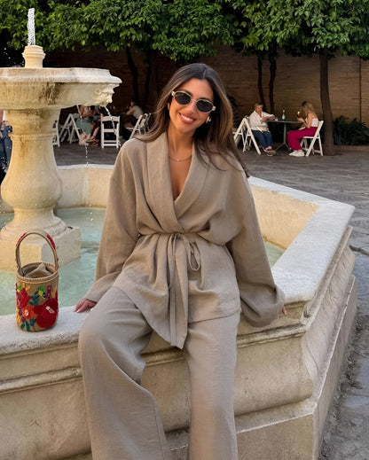 Donna sorridente con occhiali da sole, indossa abito beige elegante, seduta su una fontana in pietra. Borsa colorata accanto, ambiente esterno rilassante.