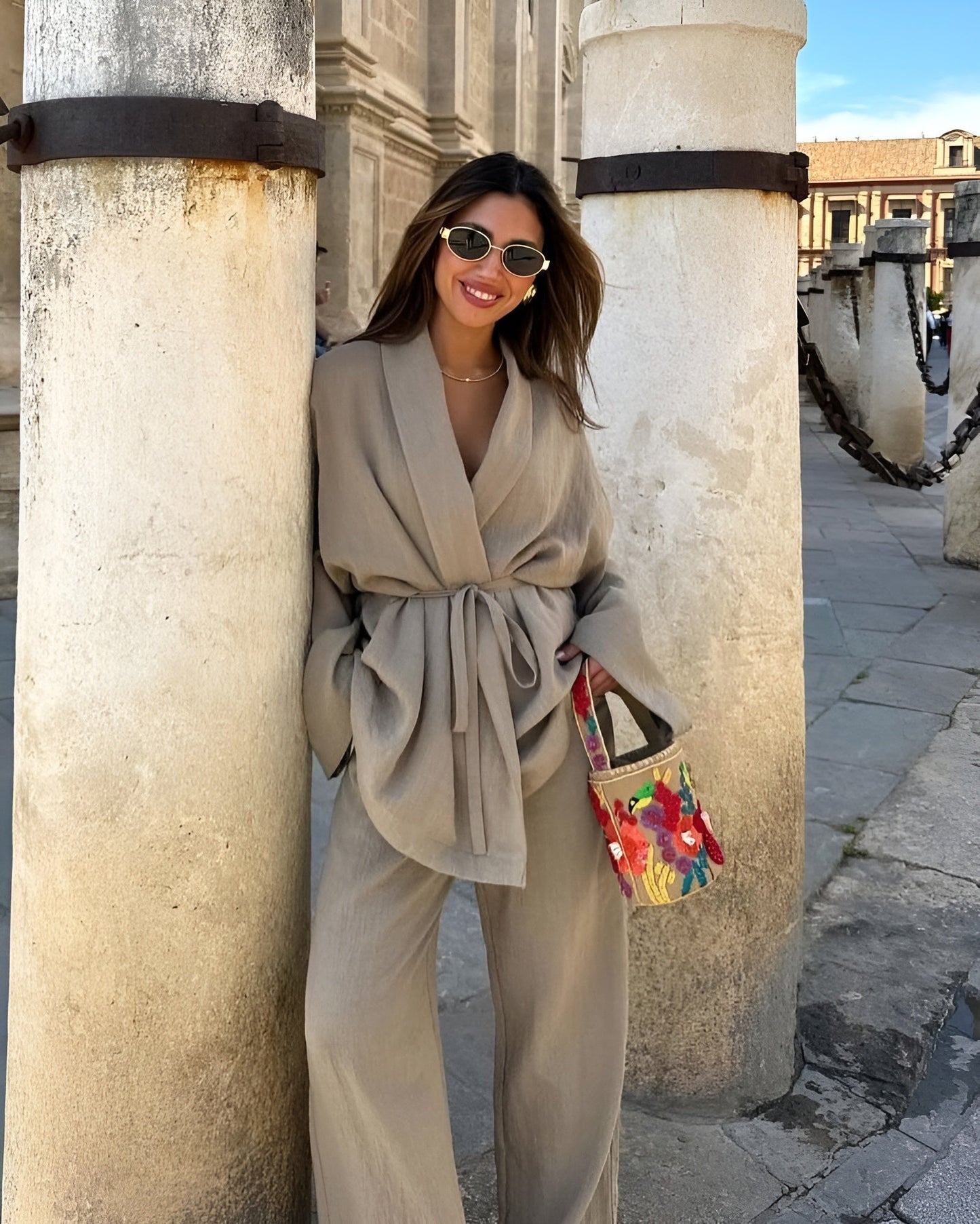 Donna sorridente con occhiali da sole indossa abito beige elegante, appoggiata a colonne storiche. Borsa colorata. Moda urbana, stile casual chic.