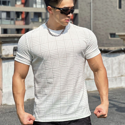 Uomo muscoloso con occhiali da sole indossa maglietta bianca a trama geometrica, posa su terrazza urbana. Moda maschile, abbigliamento estivo casual.