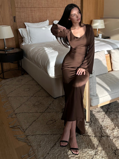 Donna in abito marrone elegante seduta su un letto in una camera moderna, illuminata da luce naturale. Arredamento minimalista, atmosfera rilassante.