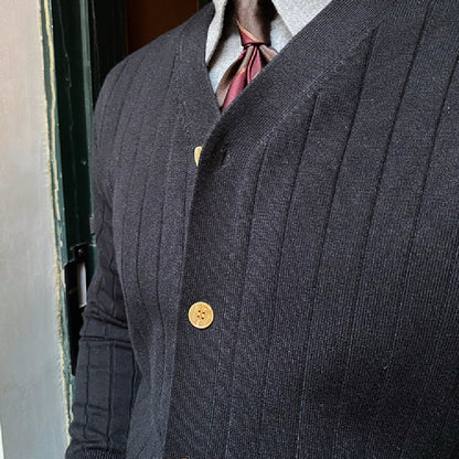 Cardigan nero a coste con bottoni dorati, indossato sopra una camicia grigia e cravatta bordeaux. Elegante abbigliamento maschile autunnale.