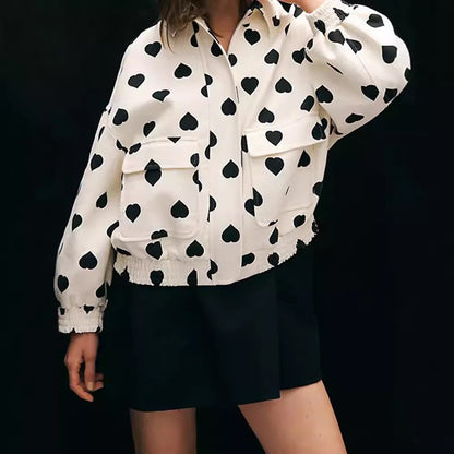 Giacca bianca con motivo a cuori neri, maniche lunghe e tasche frontali, indossata da una persona su sfondo nero. Moda autunnale elegante.