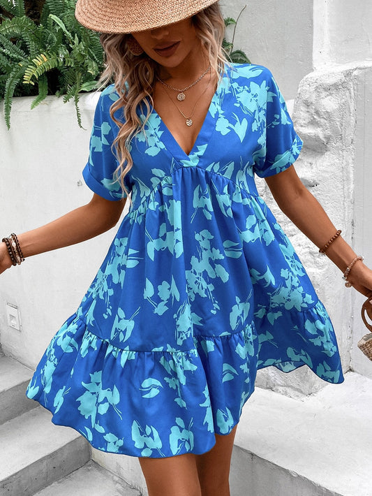 Donna con abito estivo blu a fiori, cappello di paglia, collana e bracciali, su sfondo di piante verdi. Moda estiva, abbigliamento femminile elegante.