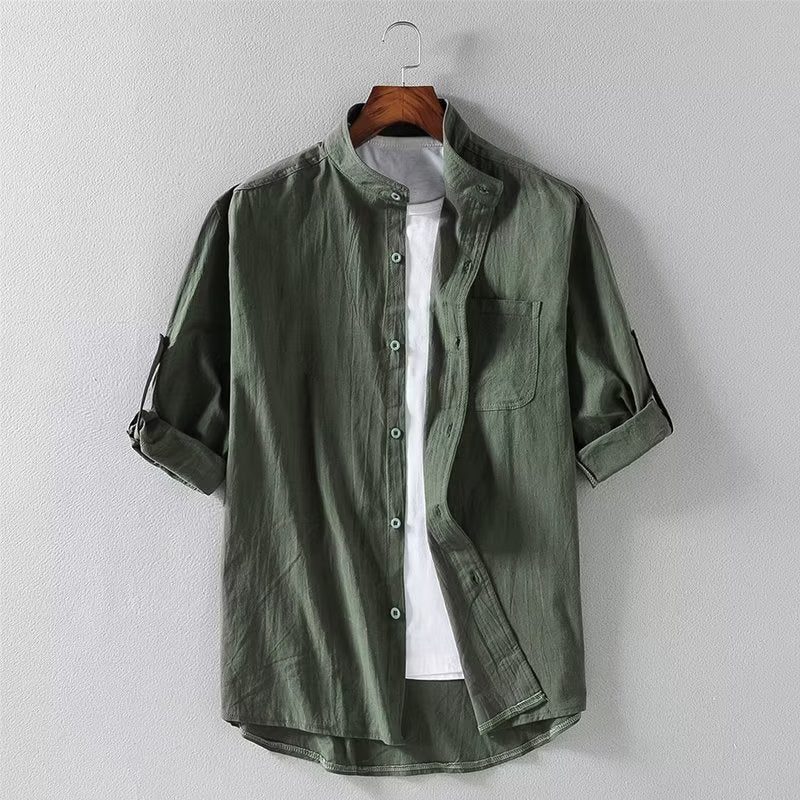 Camicia verde militare da uomo con colletto alla coreana, maniche arrotolate e taschino, appesa su gruccia. Stile casual, moda maschile.