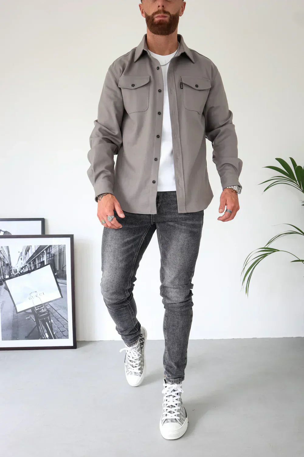 Uomo con camicia grigia e jeans neri, scarpe da ginnastica bianche, in piedi accanto a foto in bianco e nero e pianta d'appartamento.