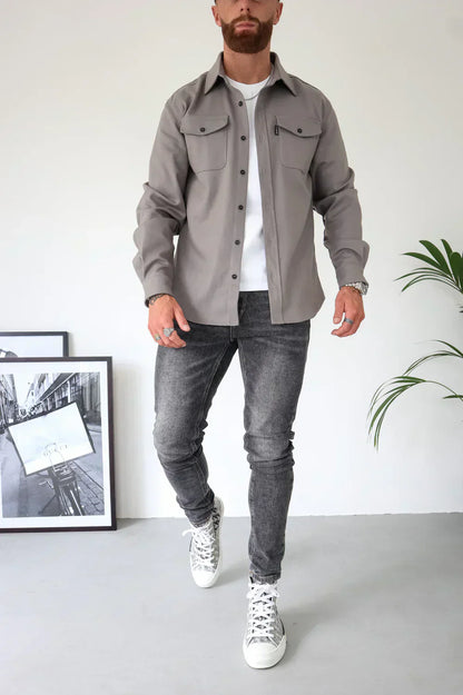 Uomo con camicia grigia e jeans neri, scarpe da ginnastica bianche, in piedi accanto a foto in bianco e nero e pianta d'appartamento.