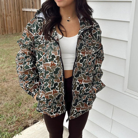 Donna con giacca mimetica verde e marrone, top bianco e pantaloni scuri, all'aperto vicino a una casa. Moda autunnale casual.