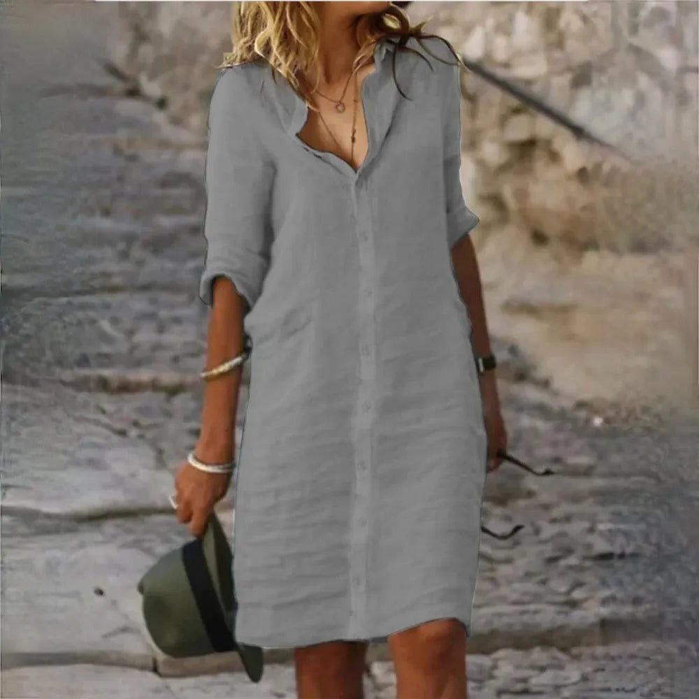 Donna con abito camicia grigio su strada acciottolata, cappello verde in mano. Moda estiva casual, look elegante e rilassato.
