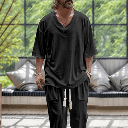 Uomo con maglietta nera oversize e pantaloni coordinati, in un ambiente moderno con cuscini e piante. Moda casual maschile, stile rilassato.