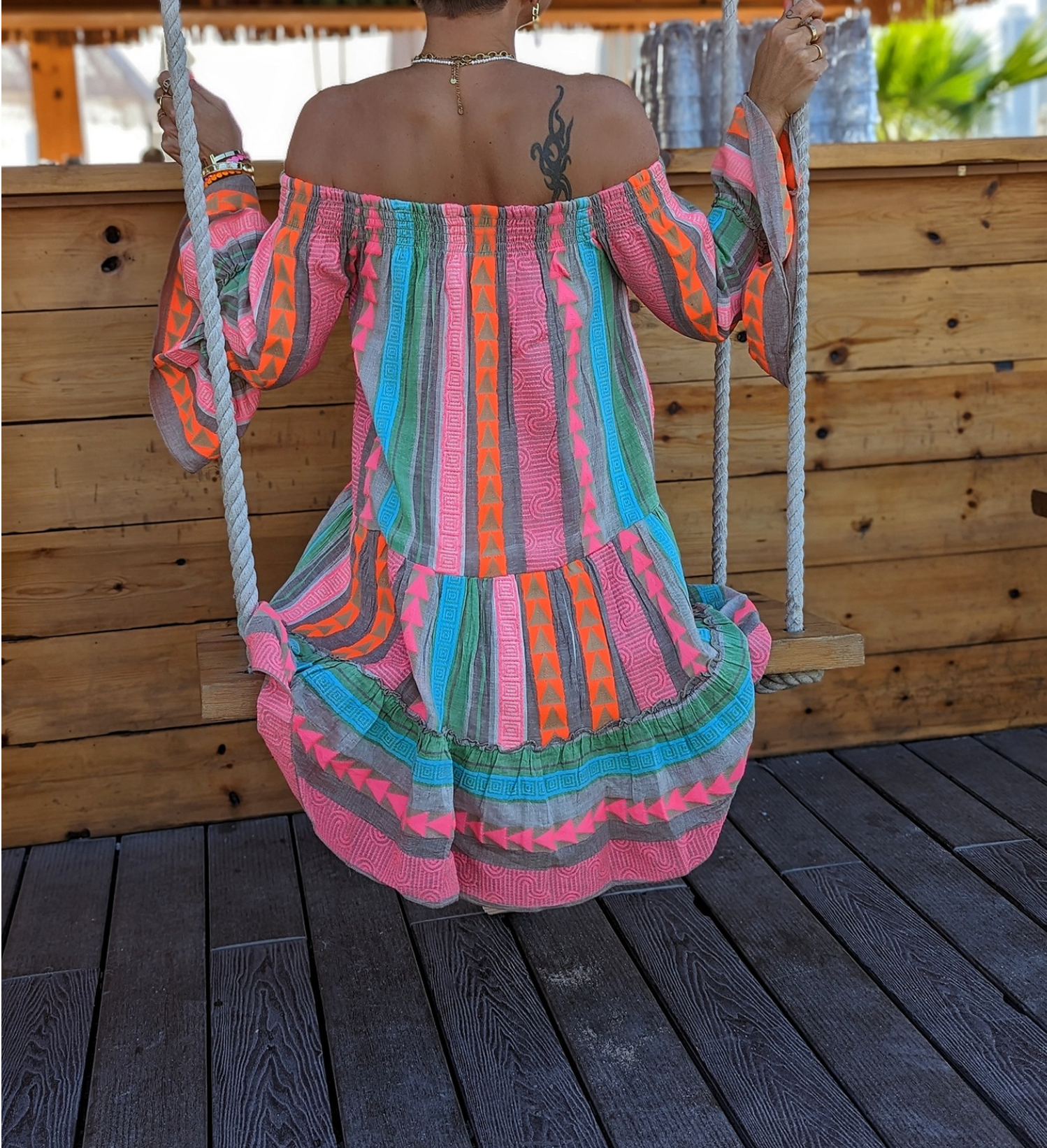 Donna su altalena in abito colorato a righe, sfondo in legno. Moda estiva, abbigliamento boho chic, relax all'aperto.