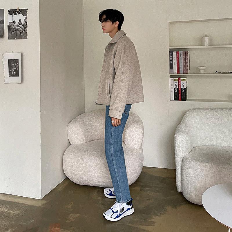 Giovane uomo in piedi in un salotto moderno, indossa giacca beige e jeans, accanto a poltrona bianca e libreria minimalista. Moda casual elegante.