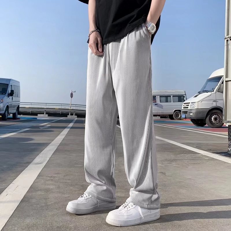 Uomo in pantaloni larghi grigi e scarpe bianche su strada urbana, sfondo con furgoni bianchi. Moda casual maschile, street style.
