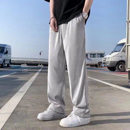 Uomo in pantaloni larghi grigi e scarpe bianche su strada urbana, sfondo con furgoni bianchi. Moda casual maschile, street style.