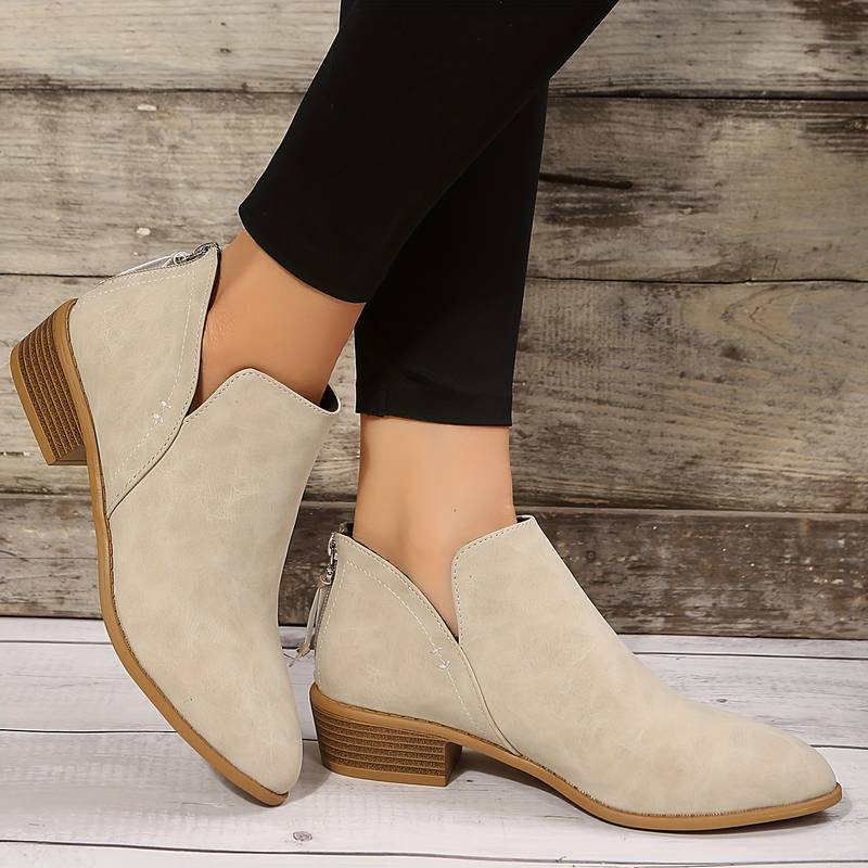 Stivaletti beige da donna in pelle scamosciata, tacco basso, chiusura con zip, indossati con leggings neri su sfondo in legno rustico.