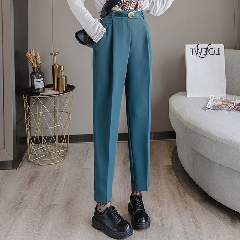 Pantaloni eleganti blu petrolio da donna, abbinati a scarpe nere e cintura dorata, su pavimento in legno chiaro. Arredamento moderno sullo sfondo.