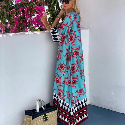 Donna in abito lungo azzurro con motivi rossi e neri, occhiali da sole, accanto a borsa di paglia e fiori rosa. Moda estiva elegante.
