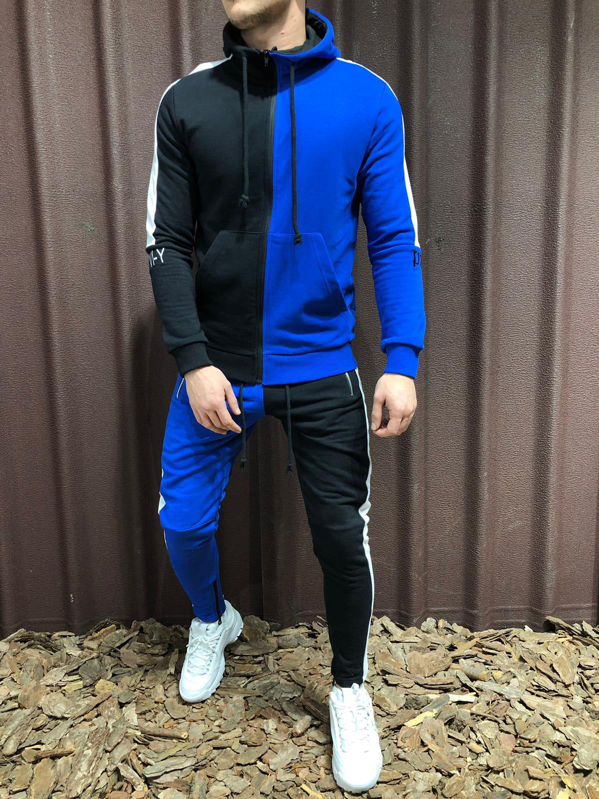 Tuta sportiva uomo bicolore blu e nera, con cappuccio e pantaloni abbinati, indossata con scarpe bianche. Moda casual, abbigliamento sportivo.
