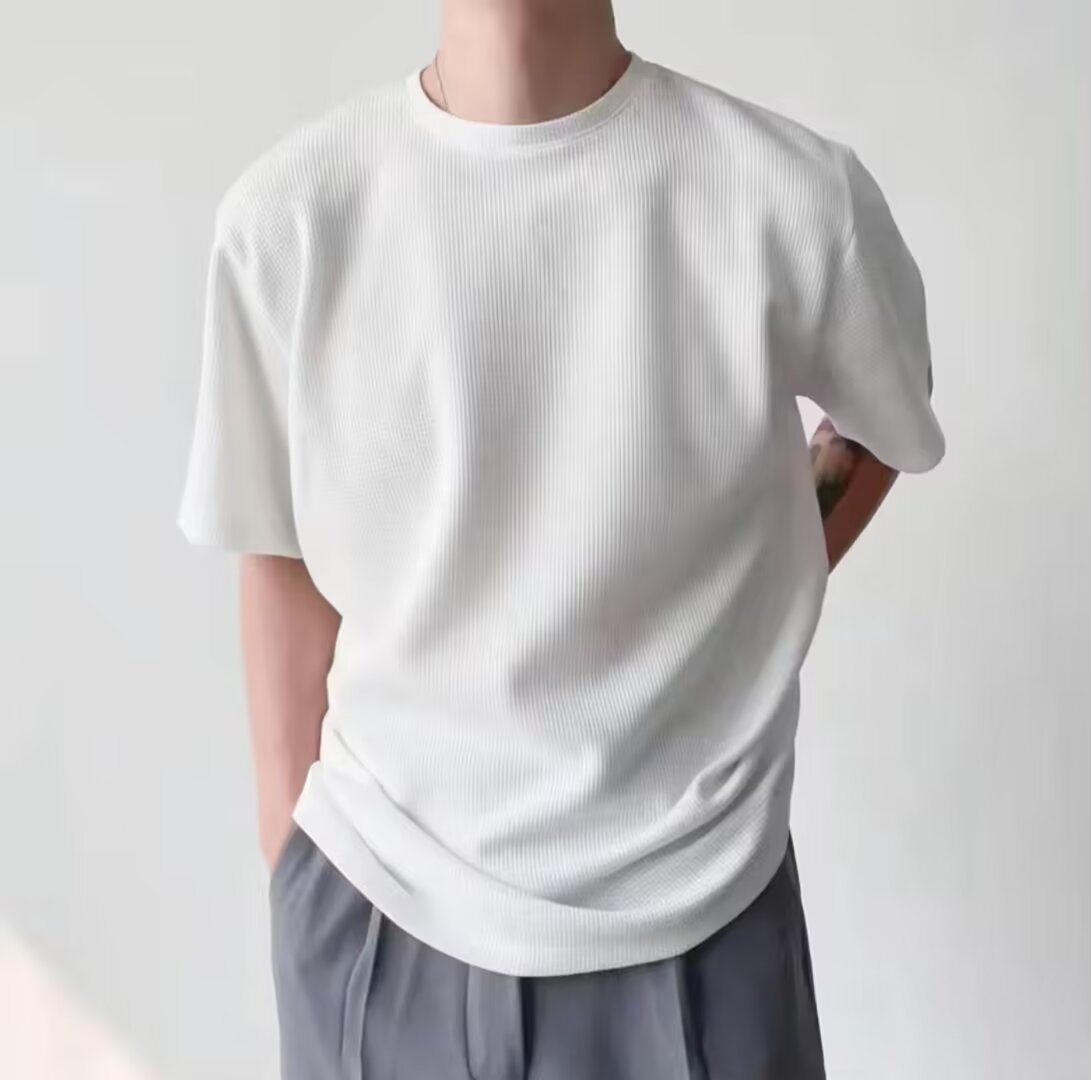 Maglietta bianca a maniche corte su uomo, tessuto a coste, abbinata a pantaloni grigi. Stile casual minimalista, moda uomo contemporanea.